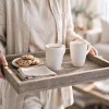 a woman holding an ottoman tray with 2 mugs of coffee and cookies